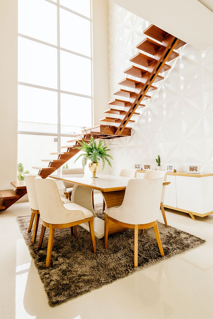 Modernes Esszimmer mit Holztisch, cremefarbenen Stühlen, goldener Vase mit grüner Pflanze. Holztreppe und großes Fenster mit weißer geometrischer Wand.