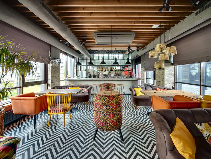 Stylish cafe interior with geometric floor, colorful chairs, and wooden ceiling. Large windows and hanging lights create a cozy atmosphere.